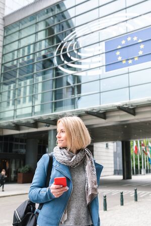 A young woman stands with a telephone in her hands opposite the European Parliament building in Brussels, Belgium.のeditorial素材