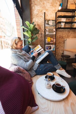 Young woman tourist sitting in a cafe by the window, reading a magazine and drinking coffeeのeditorial素材