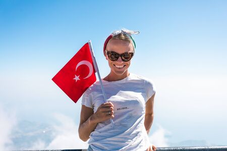 Young woman tourist in denim shorts and a white T-shirt stands on a viewing platform of Tahtali Mountain in Kemer with a red Turkey flag in handsの写真素材