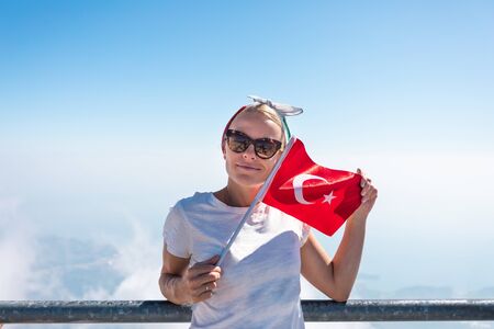 Young woman tourist in denim shorts and a white T-shirt stands on a viewing platform of Tahtali Mountain in Kemer with a red Turkey flag in handsの写真素材