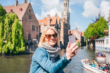 Beautiful young girl takes selfie photo of the famous tourist destination with a canal in Bruges, Belgiumの写真素材