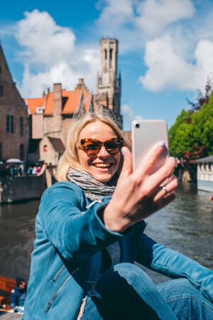 Beautiful young girl takes selfie photo of the famous tourist destination with a canal in Bruges, Belgiumの写真素材