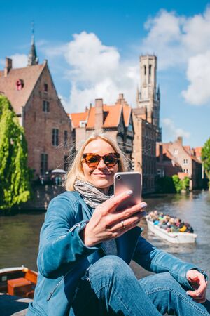 Beautiful young girl takes selfie photo of the famous tourist destination with a canal in Bruges, Belgiumの写真素材