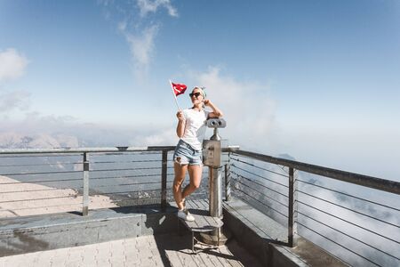 Young woman tourist in denim shorts and a white T-shirt stands on a viewing platform of Tahtali Mountain in Kemer with a red Turkey flag in her hands near binoculars against the backdrop of a beautiful panorama of mountains, Turkeyの写真素材
