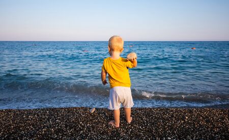 A little boy stands in the background of the blue sea and throws stones into the water at sunsetの写真素材
