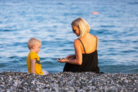 Mother and son throw pebbles into the water on the beach at sunsetの写真素材
