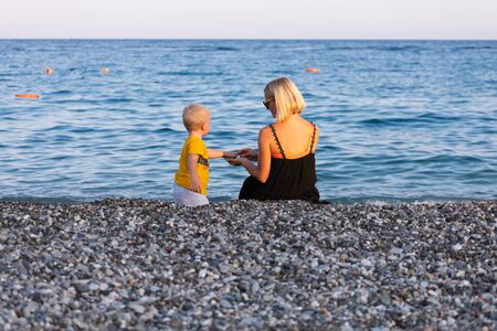 Mother and her little son throw pebbles into the water on the beach at sunset during their vacationの写真素材