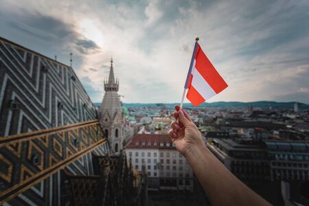 A woman holds in her hand the flag of Austria on a viewing platform of St. Stephen's Cathedral in Vienna against the backdrop of a beautiful sunset sky and city panorama.の写真素材