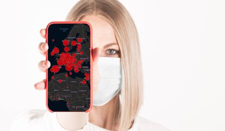 Portrait of a woman in a medical mask with a red phone in her hands with a map on the phone with the epicenters of the disease isolated on whiteの写真素材