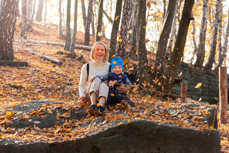 Boy and his mother are sitting in the autumn forest and tossing leaves.の写真素材