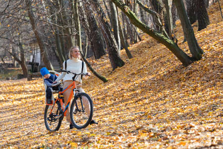 Happy mother leads a bicycle with a child strapped in the back in the autumn park.の写真素材