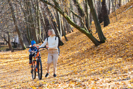 Happy mother leads a bicycle with a child strapped in the back in the autumn park.の写真素材