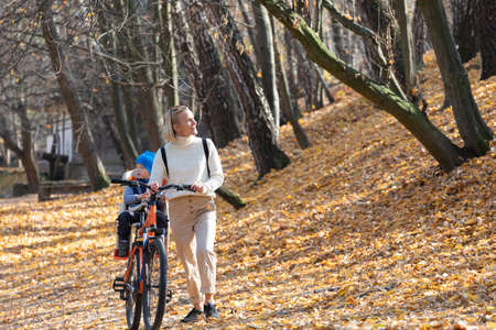 Happy mother leads a bicycle with a child strapped in the back in the autumn park.の写真素材