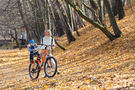 Happy mother leads a bicycle with a child strapped in the back in the autumn park.の写真素材