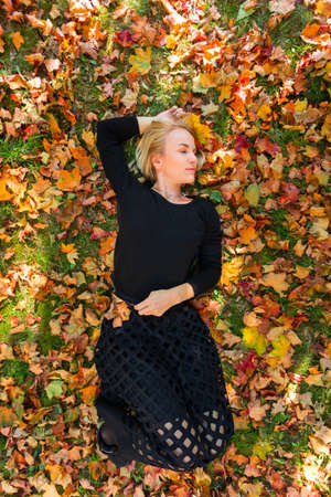 A young woman lies on fallen orange leaves and holds yellow maple leaves in her hand.の写真素材