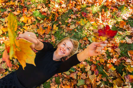 A young woman lies on fallen orange leaves and holds yellow maple leaves in her hand.