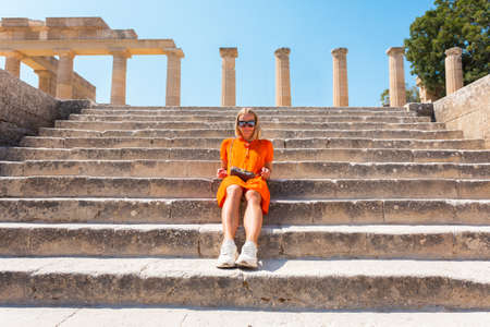 Young happy blonde woman sits on the steps of the Acropolis in the city of Lindos, Rhodes, Greeceの写真素材