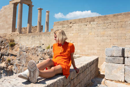 A young blonde woman is sitting and enjoying the view of the coast near the city of Lindos, Greece.の写真素材