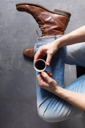 Men's hand holding cup of coffee and leg in jeans with old travel leather boots shoes. Male fashion life style conceptの写真素材