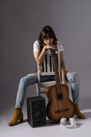 Young woman holdilng classic guitar sitting on a chair. Musician in studio near acoustic guitar and music columnの写真素材