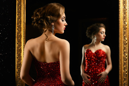 fashionable woman standing in front of gold mirror in red gownの写真素材
