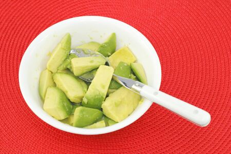 Close up of a spoon in a white bowl of just avocado pieces mixed with lime juice on a red placemat.の写真素材
