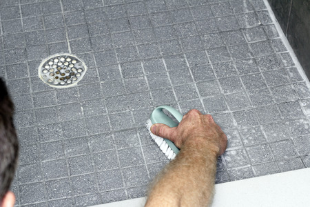 Male leaning into a shower, scrubbing a tile floor with a scrub brush and soap. Gray tile floor with...