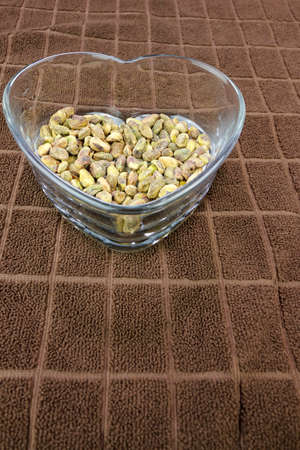 Roasted shell-less pistachios with sea salt in a heart shaped clear glass dish on a brown towel with rectangle pattern. On a brown towel is a heart shaped clear glass dish with salted pistachios foodの写真素材