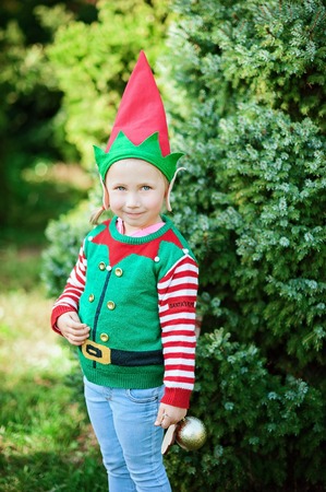 Little girl in elf sweater and hat waiting for a Christmas in the wood. Half-length portrait of a little child near the christmas treeの写真素材