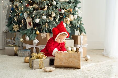 The little girl under the Christmas tree. baby girl in Santa Claus hat with gifts under Christmas tree at fireplaceの写真素材