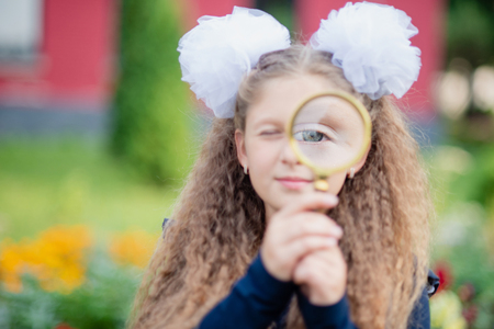 Portrait of a beautiful young Schoolgirl looking through a magnifying glass and sitting at a desk on the background autumn park. Farewell Bell. day of knowledge. beginning of the school year. school equipment pencils books glass desk.の写真素材