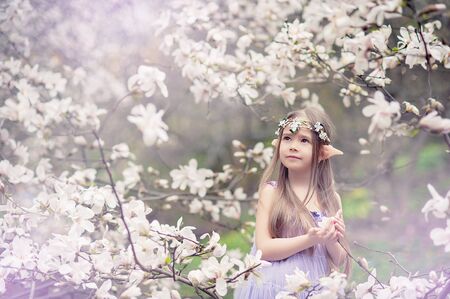 fairy tale girl. Portrait of mystic elf child. Cosplay character. Portrait of an elf in a blooming magnolia garden. A girl with long ears touches the flowers of white magnoliaの写真素材
