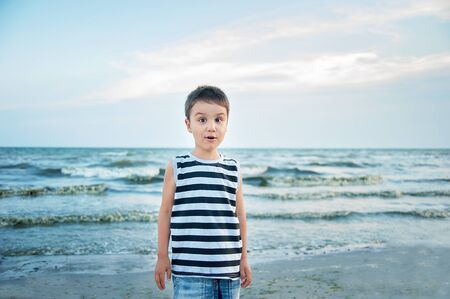Wondered amazed cute boy in a striped T-shirt at the seaside questioned surprised. Summer vacation. happy kid playing on beach at the sunset timeの写真素材