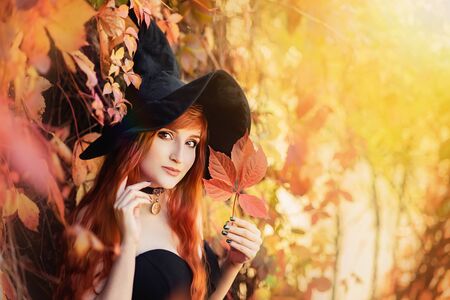 Halloween Beautiful young Witch girl in witches hat on autumn magic forest background. Portrait of model girl with autumn bright leaves, Fall look style. redhead girl with big eyes magical sightの写真素材