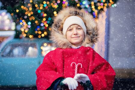 Child on christmas market. cute lovely girl in red hat standing at street and drinking tea or cacao, hot coffee outdoors in winter snow day. Christmas lightsの写真素材