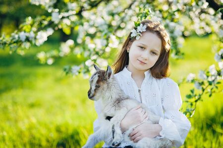Smiley girl with a little goat sitting in the green grass in a flowering apple gardenの写真素材