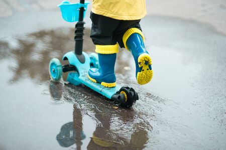 Happy little kid in a yellow raincoat rides a scooter across the puddle on warm summer day after rain. Active leisure for children. Child having fun outdoors.の写真素材
