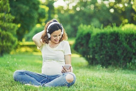 pregnant girl in headphones outdoors. Happy young pregnant woman sitting at park fun greeting online...