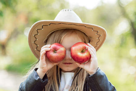 portrait of girl eating red organic apple outdoor. Harvest Concept. Child picking apples on farm in autumn. Children and Ecology. Healthy nutrition Garden Food. Girl holding in front of her face appleの写真素材