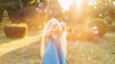 Child whirling, dancing plays on the meadow. Girl having fun with bubbles. Cute little longhair blonde girl dancing with soap bubbles at sunset park.