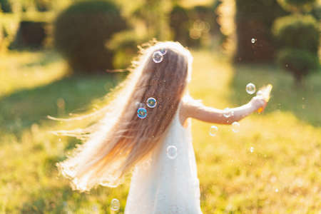 Child whirling, dancing plays on the meadow. Girl having fun with bubbles. Cute little longhair blonde girl dancing with soap bubbles at sunset park.の写真素材