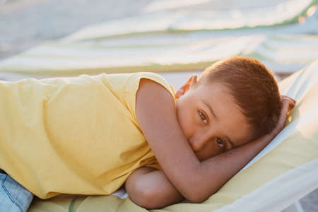 boy in yellow t-shirtsitting on bench near sea alone on beach at sunset. Happy family travel. Summer vacation with child.の写真素材