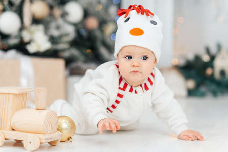 Happy Holidays, New year. Little child under Christmas tree. baby boy in snowman hat with gifts under Christmas tree with many gift boxes presents. Cozy warm winter evening at home. Xmas timeの写真素材
