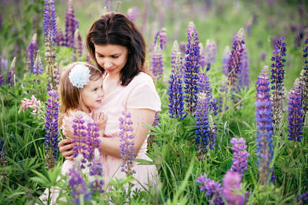 Happy family outdoor. Middle-aged mother hugs her daughter in field. Mother's Day. Portrait of smiling beautiful young woman and small child in her arms sitting on grass, maternity concept.の写真素材
