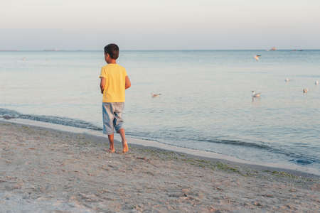 boy in a yellow shirt looks far away to horizon of sea standing alone on beach at sunset. View from back. Happy family travel. Summer vacation with child.の写真素材