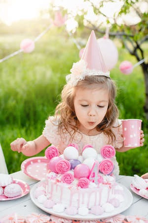 Happy birthday little girl making wish blowing candles on cake with pink decor in beautiful garden. 4 years birthday. child eat happy birthday pink cupcake. colorful pastel decoration outdoorの写真素材
