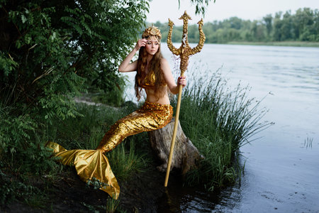 Fantasy woman real mermaid with trident myth goddess of sea with golden tail sitting in sunset on rocks .. Gold hair crown shells pearls jewelry. Mermaid sitting on shore. fantasy concept.の写真素材