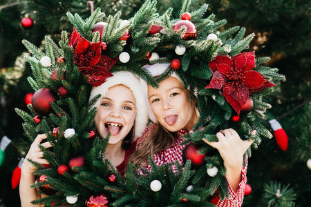 Merry Christmas. Portrait of two happy funny children girls in Santa hat with Christmas wreath. Happy Holidays. Fairy Magic. Happy kids enjoying holiday.の写真素材