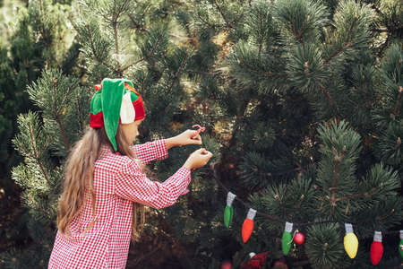 Child waiting for Christmas in wood in summer. portrait of little girl in red dress decorating christmas tree. winter holidays and people concept. Merry Christmas and Happy Holidaysの写真素材
