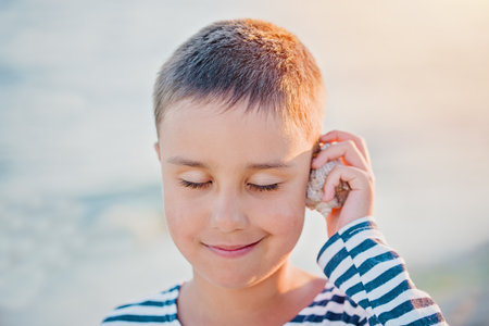 Cute little boy listening sound of sea in shell at the beach. Child in striped T-shirt at seaside. Summer vacation in Ukraine Koblebo. happy kid playing on beach at sunset time. Happy childhoodの写真素材